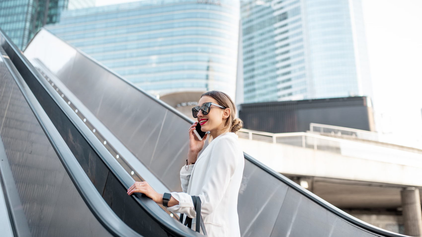 Stylish businesswoman in white suit talking phone while going up on the escalator at the business centre outdoors in Paris