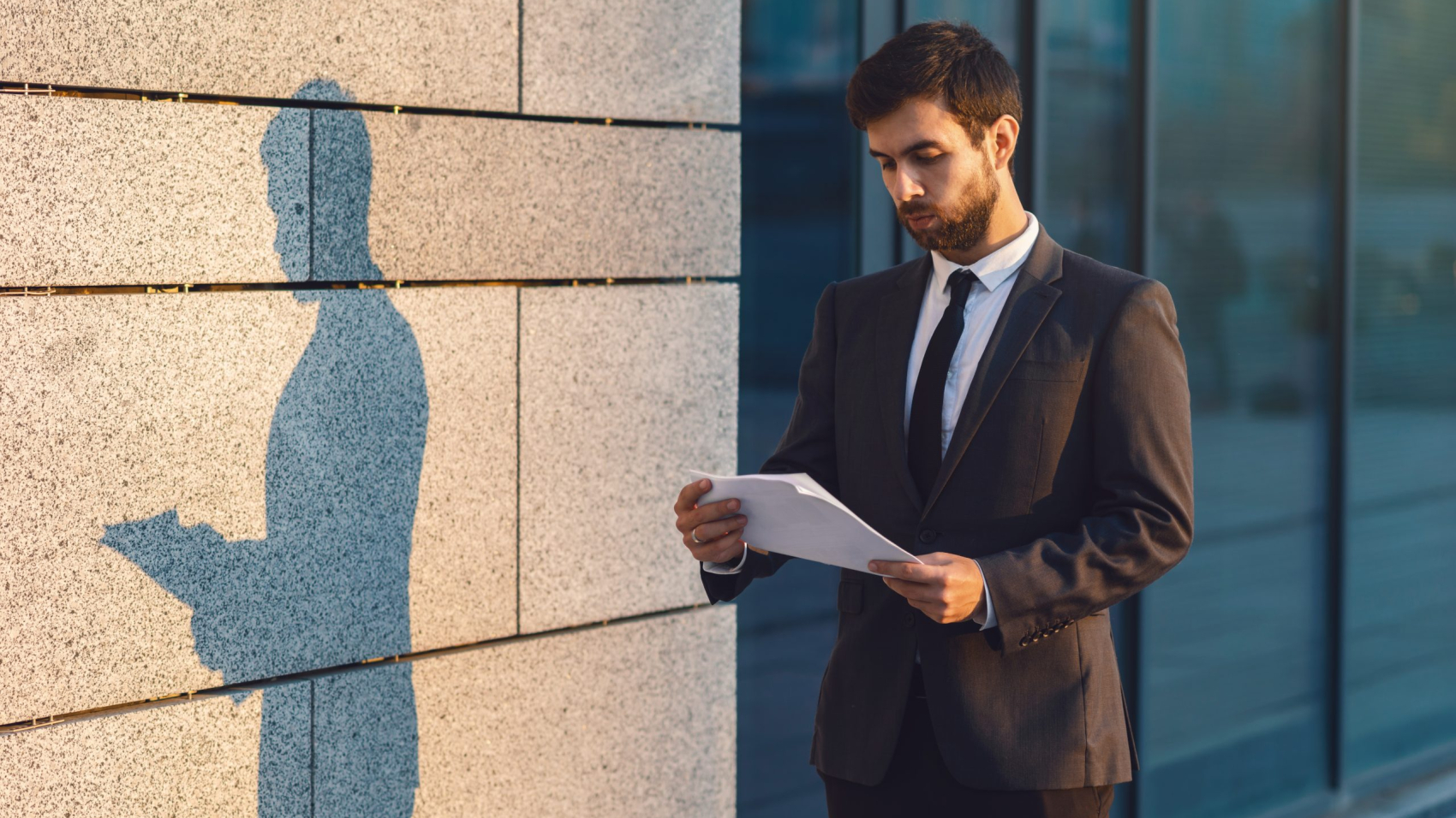 Serious young businessman in a classic suit reading advantageous business document in paper form. Standing outdoors near office building under the sunshine. Cast a shadow